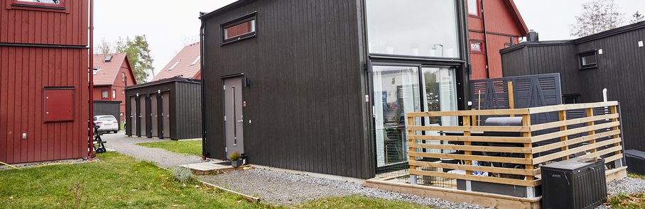 Svart attefallshus med stora fönster och trädäck. Attefallshuset ligger på samma tomt som parhus i Väsjön.