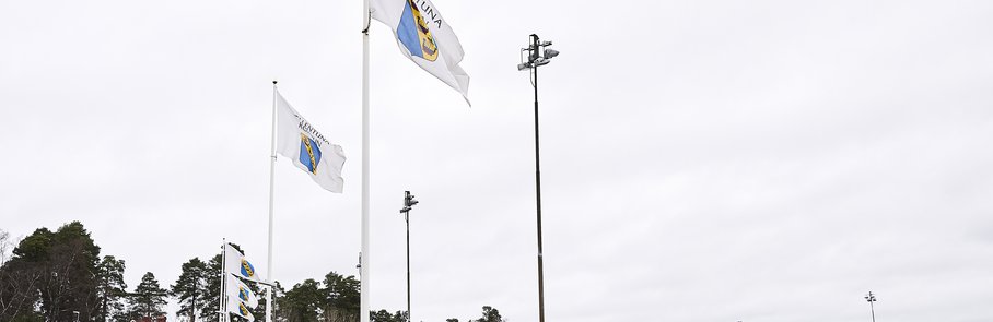 Flaggor som vajar i vinden vid idrottsanläggningen Sollentunavallen. Flaggorna är vita  med Sollentuna kommuns logga i färg.
