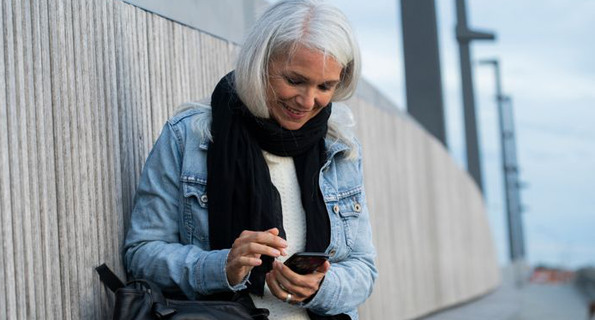 Gråhårig kvinna sitter vid betongvägg och använder sin mobiltelefon.