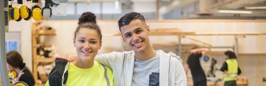 Full length portrait of smiling female trainee standing with arm around male coworker at workbench