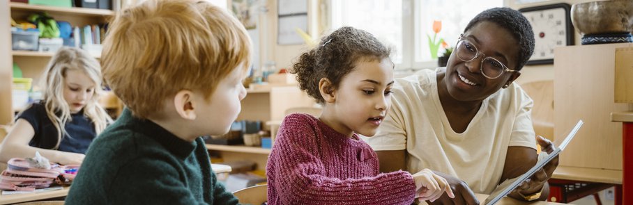 Smiling teacher assisting boy and girl with digital tablet sitting at desk in classroom